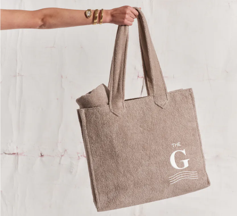 A woman's hand wearing a gold bracelate holding a luxury tote, light brown, bag with the brand name G wich is written with white letters.